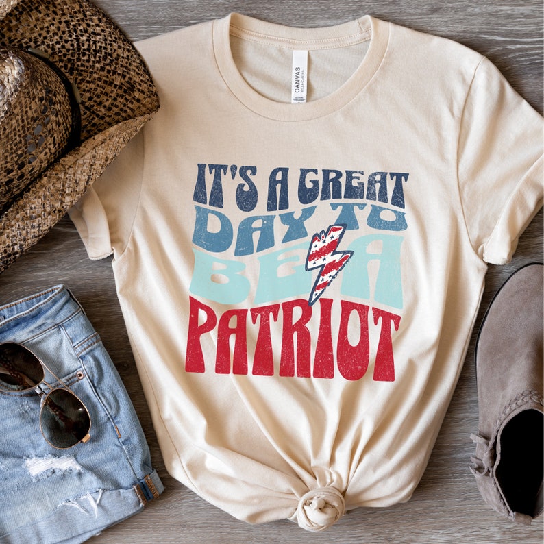 May include: A beige t-shirt with the text "It's a great day to be a patriot" in red, blue and white with a lightning bolt graphic. The text is in a retro font style.