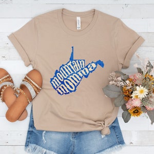 May include: A tan t-shirt with a blue outline of the state of West Virginia. The words "mountain momma" are written in a yellow font inside the state outline.