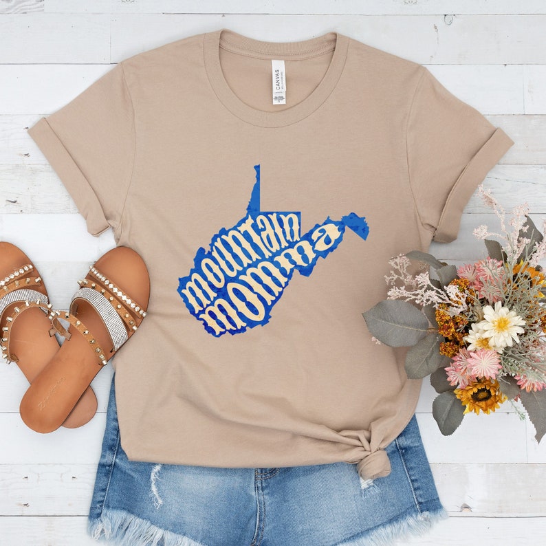 May include: A tan t-shirt with a blue outline of the state of West Virginia. The words "mountain momma" are written in a yellow font inside the state outline.