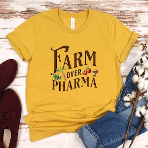 May include: A mustard yellow t-shirt with the text "FARM OVER PHARMA" in brown lettering. The text is surrounded by small images of vegetables and a mushroom.