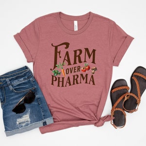 May include: A mauve t-shirt with the text "FARM OVER PHARMA" in brown lettering. The text is surrounded by images of vegetables and a mushroom.