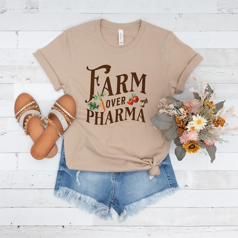 May include: A tan t-shirt with the text "FARM OVER PHARMA" in brown lettering. The text is surrounded by small images of vegetables and a mushroom.