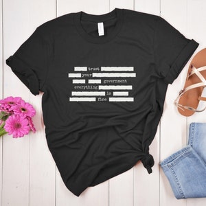 Op de afbeelding: Zwart T-shirt met witte tekst die "trust your government everything is fine" luidt. De tekst is in een typemachinelettertype en wordt gescheiden door witte lijnen.