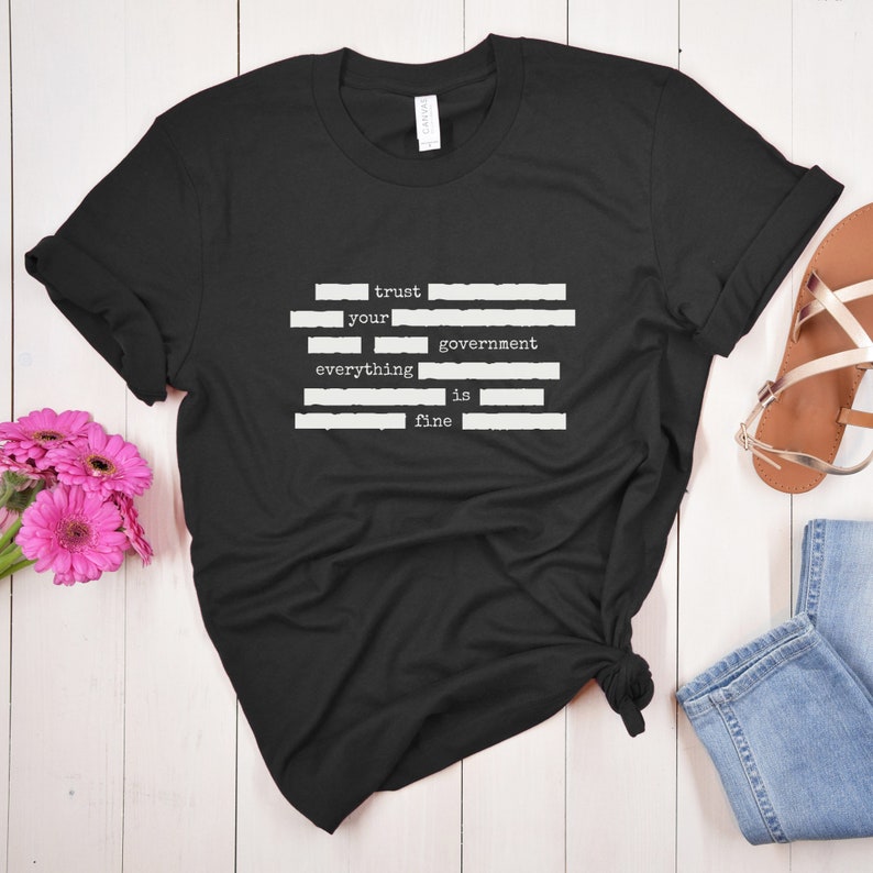 Op de afbeelding: Zwart T-shirt met witte tekst die "trust your government everything is fine" luidt. De tekst is in een typemachinelettertype en wordt gescheiden door witte lijnen.