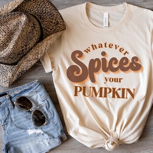 May include: A cream-coloured t-shirt with the text "whatever Spices your PUMPKIN" in brown and orange lettering. The shirt is styled with denim shorts, sunglasses, and a straw hat.