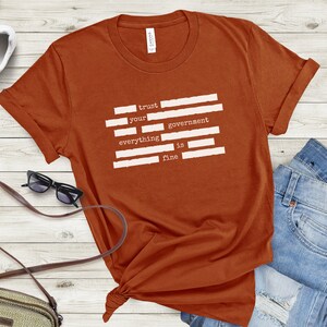 Op de afbeelding: Een roestkleurig T-shirt met een witte afbeelding waarop staat "trust your government everything is fine". De afbeelding bestaat uit witte balken die lijken op een Amerikaanse vlag.