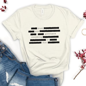 Op de afbeelding: Een wit T-shirt met zwarte tekst die "trust your government everything is fine" luidt. De tekst is in een typemachinelettertype en is gerangschikt in een verticale kolom.