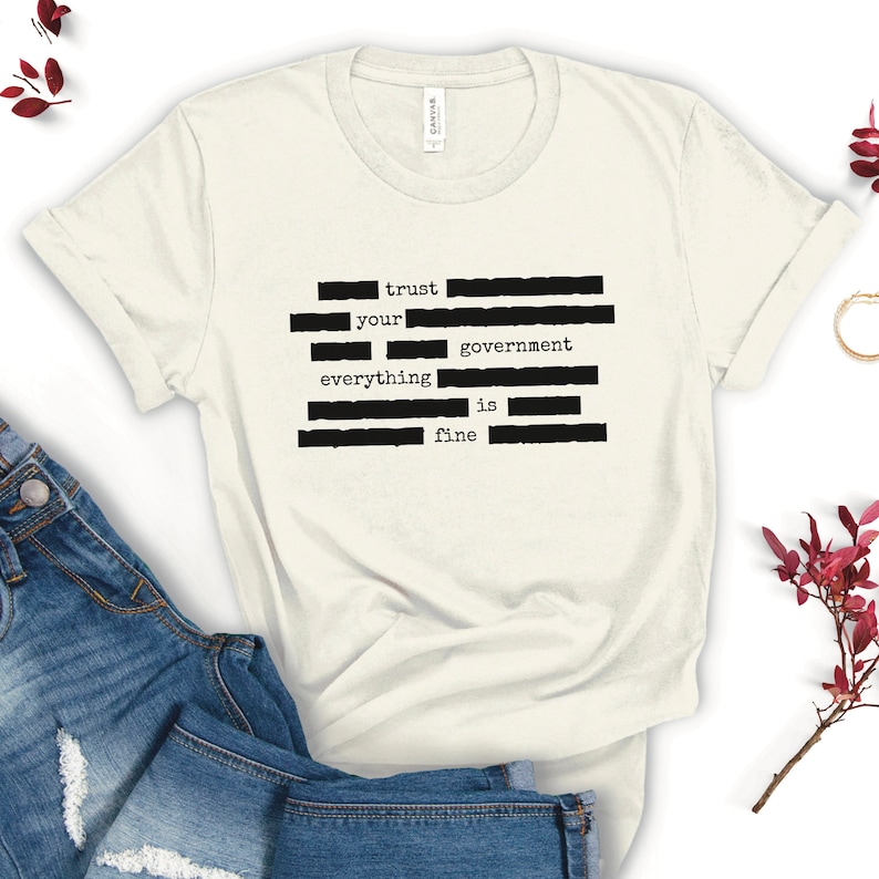Op de afbeelding: Een wit T-shirt met zwarte tekst die "trust your government everything is fine" luidt. De tekst is in een typemachinelettertype en is gerangschikt in een verticale kolom.