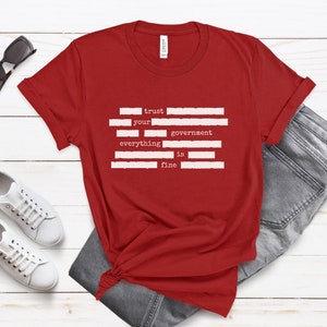 Op de afbeelding: Rood T-shirt met witte tekst die "trust your government everything is fine" luidt. De tekst is in een typemachinelettertype en is verticaal gerangschikt.