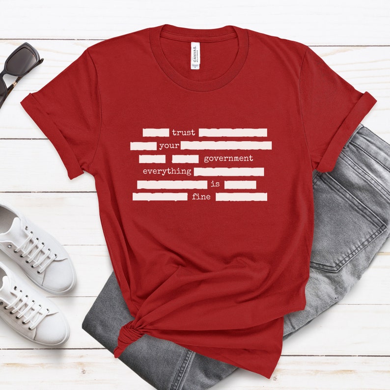 Op de afbeelding: Rood T-shirt met witte tekst die "trust your government everything is fine" luidt. De tekst is in een typemachinelettertype en is verticaal gerangschikt.
