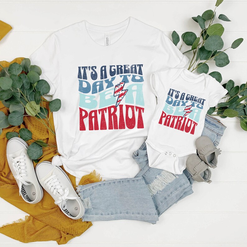 May include: Two white t-shirts with the text "It's a great day to be a patriot" in red, blue, and white with a lightning bolt graphic. The shirts are on a white surface with blue jeans, white trainers, and a yellow scarf.