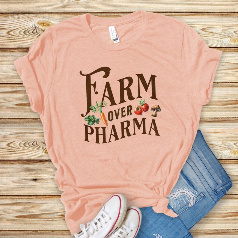 May include: A peach-colored t-shirt with the text "FARM OVER PHARMA" in brown lettering. The text is surrounded by small images of vegetables and mushrooms.