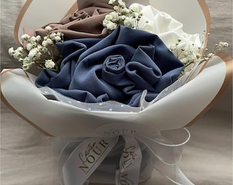 Può includere: Un bouquet di rose in tessuto nei toni del blu, marrone e bianco, accentuato da piccoli fiori bianchi di gipsofila. Il bouquet è avvolto in carta bianca e marrone chiaro, legato con un nastro bianco stampato con il testo Collection NOUR.