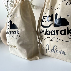 May include: Two white canvas drawstring bags with black and gold lettering. The bags have a mosque design and the words "Eid Mubarak" on them. One bag says "Adem" and the other says "Ossia".