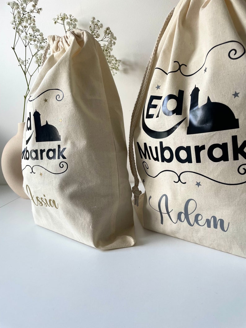 May include: Two white canvas drawstring bags with black and gold lettering. The bags have a mosque design and the words "Eid Mubarak" on them. One bag says "Adem" and the other says "Ossia".
