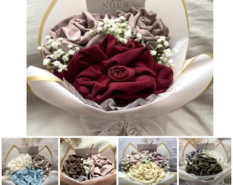 Pode incluir: Um buquê de flores de tecido dispostas como rosas em tons de bordô e malva, acentuado com pequenas flores brancas de gipsófila. O buquê é embrulhado em papel branco com borda dourada e inclui um cartão com a etiqueta Collection NOUR.