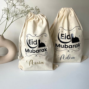 May include: Two white cotton drawstring bags with black text and a black silhouette of a mosque. The text on the bags reads "Eid Mubarak" with the names "Assia" and "Adem" written below.