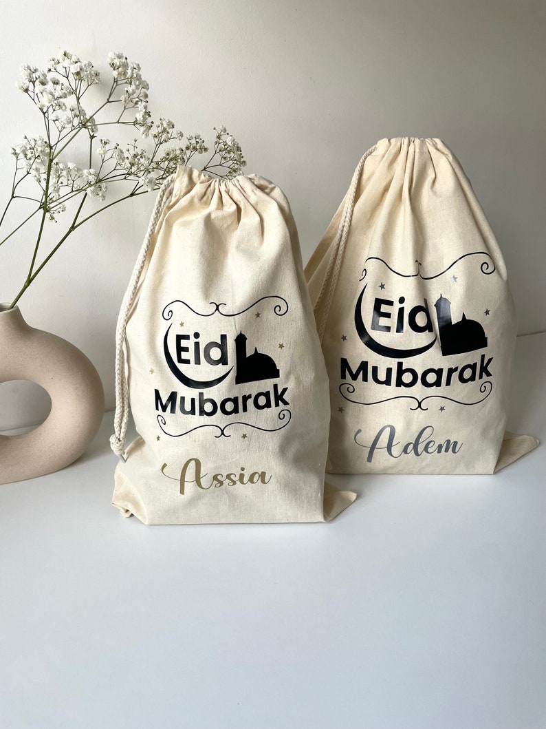 May include: Two white cotton drawstring bags with black text and a black silhouette of a mosque. The text on the bags reads "Eid Mubarak" with the names "Assia" and "Adem" written below.