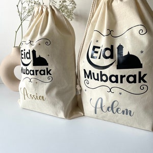 May include: Two white drawstring bags with black text that reads "Eid Mubarak" and a mosque silhouette. The bag on the left has the name "Assia" in gold lettering and the bag on the right has the name "Adem" in silver lettering.