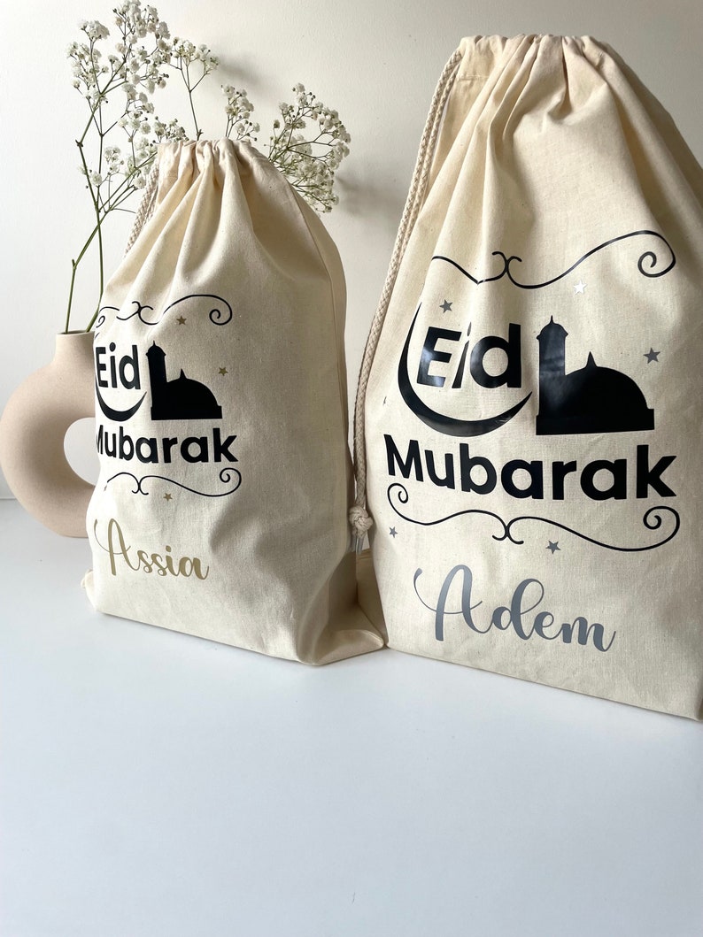 May include: Two white drawstring bags with black text that reads "Eid Mubarak" and a mosque silhouette. The bag on the left has the name "Assia" in gold lettering and the bag on the right has the name "Adem" in silver lettering.