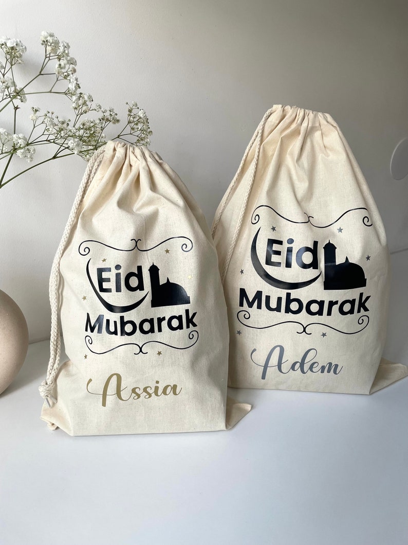 May include: Two white canvas drawstring bags with black text that reads "Eid Mubarak" and a silhouette of a mosque. One bag has the name "Assia" printed in gold below the text and the other bag has the name "Adem" printed in silver below the text.