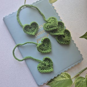 May include: Green crocheted heart-shaped bookmarks and embellishments. The hearts are attached to thin, crocheted stems. The items are arranged on a light blue notebook with the words "protect your peace" visible. Green leaves are in the background.