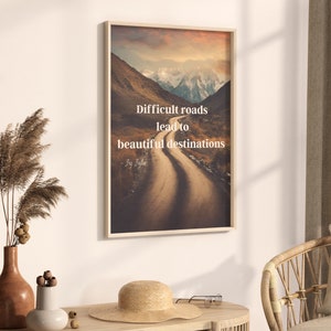 Puede incluir: Una impresión enmarcada con una cita motivacional: "Los caminos difíciles conducen a hermosos destinos." La imagen muestra un camino de tierra sinuoso que conduce a través de un valle de montaña con picos cubiertos de nieve en la distancia.