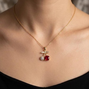Peut inclure: Un collier doré avec un pendentif cerise. Le pendentif présente une cerise en pierre rouge et une cerise blanche ornée de cristaux. Le collier est porté par une personne.