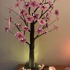 Puede incluir: Un árbol decorativo con tronco y ramas marrones, adornado con flores rosas de ganchillo. Las flores están iluminadas con pequeñas luces blancas, creando un brillo cálido. El árbol se asienta sobre una base cuadrada.
