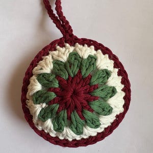 Op de afbeelding: Een ronde, gehaakte onderzetter met een lus om op te hangen. De onderzetter heeft een centraal bordeauxrood bloemmotief, groene en witte bloemblaadjes en een bordeauxrode rand. De lus is ook bordeauxrood.