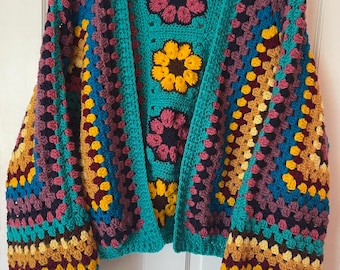 May include: A teal blue crocheted cardigan with colorful granny square patterns. The cardigan has a V-neck and long sleeves with a puff sleeve design.
