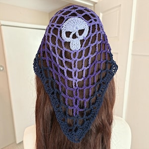 May include: A triangular crochet headscarf featuring an open net pattern that transitions from a light purple at the top to a deep navy blue at the bottom. A light purple crocheted skull motif is centered near the top edge of the headscarf.