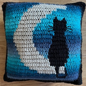 May include: A crocheted pillow with a black cat silhouette and a crescent moon design. The pillow is made with a gradient of blue yarn, with a white crescent moon and a black cat silhouette.