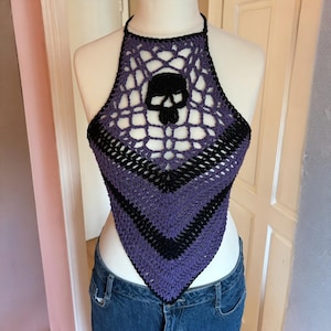 Puede incluir: Top halter de ganchillo hecho a mano en tonos morado y negro. El top presenta un diseño de calavera en el centro y un patrón geométrico. El top halter se muestra en un maniquí, mostrando su diseño único y artesanía.