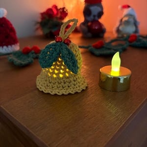 May include: A crocheted bell-shaped ornament with green leaves and red berries, illuminated from within. A small, battery-operated candle sits nearby. Other Christmas decorations are in the background, including a Santa hat and a gnome.