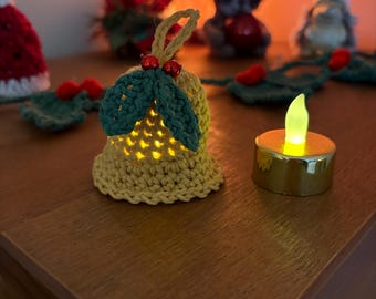 May include: A crocheted bell-shaped ornament with green leaves and red berries, illuminated from within. A small, battery-operated candle sits nearby. Other Christmas decorations are in the background, including a Santa hat and a gnome.
