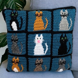 May include: A teal blue crocheted pillow with a square pattern featuring nine different colored cats. The cats are crocheted in black, white, gray, and orange.