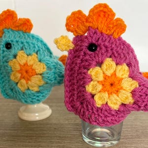 Può includere: Due ornamenti a forma di uccello fatti a mano all'uncinetto. Uno è turchese con accenti arancioni e gialli, l'altro è magenta con accenti arancioni e gialli. Entrambi hanno occhi neri e sono appoggiati su un supporto bianco e un bicchiere.