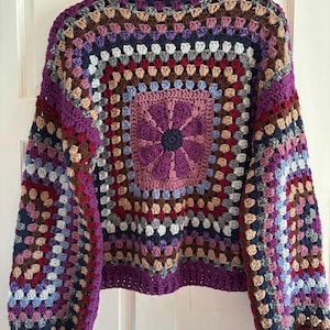 Könnte beinhalten: Ein mehrfarbiger Häkelpullover mit einem Blumendesign in der Mitte. Der Pullover hat einen hohen Kragen und lange Ärmel.