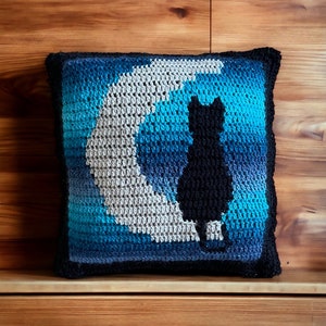 May include: A crocheted pillow with a blue, gray, and white ombre design featuring a black cat silhouette sitting on a crescent moon.