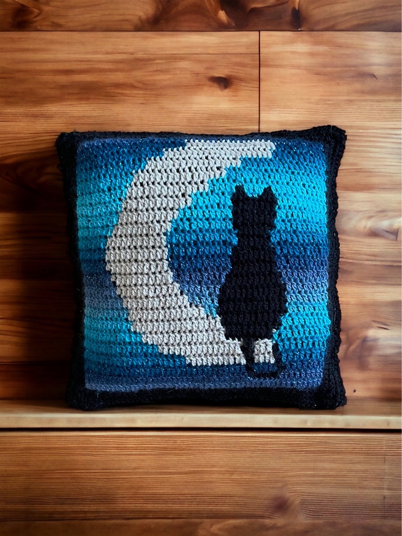 May include: A crocheted pillow with a blue, gray, and white ombre design featuring a black cat silhouette sitting on a crescent moon.