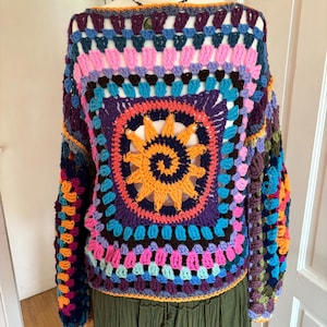 Puede incluir: Un suéter de crochet colorido con un gran diseño circular multicolor en el centro. El suéter tiene mangas largas y un ajuste holgado.