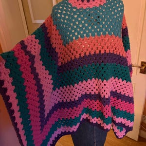Puede incluir: Un poncho de ganchillo hecho a mano y colorido en tonos turquesa, rosa, morado y coral. El poncho presenta un cuello alto acanalado y un diseño geométrico calado. La prenda se exhibe en un maniquí.