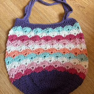 May include: A handmade crochet tote bag in a vibrant array of colors. The bag features a purple base and handle, with alternating rows of white, pink, orange, and teal. The bag is designed with a shell stitch pattern.