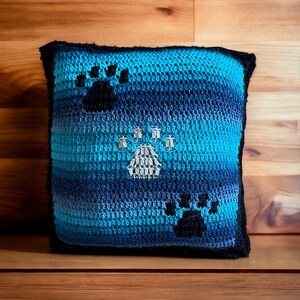 May include: A blue and black crocheted pillow with paw print designs. The pillow is made with a gradient yarn that transitions from dark blue to light blue.