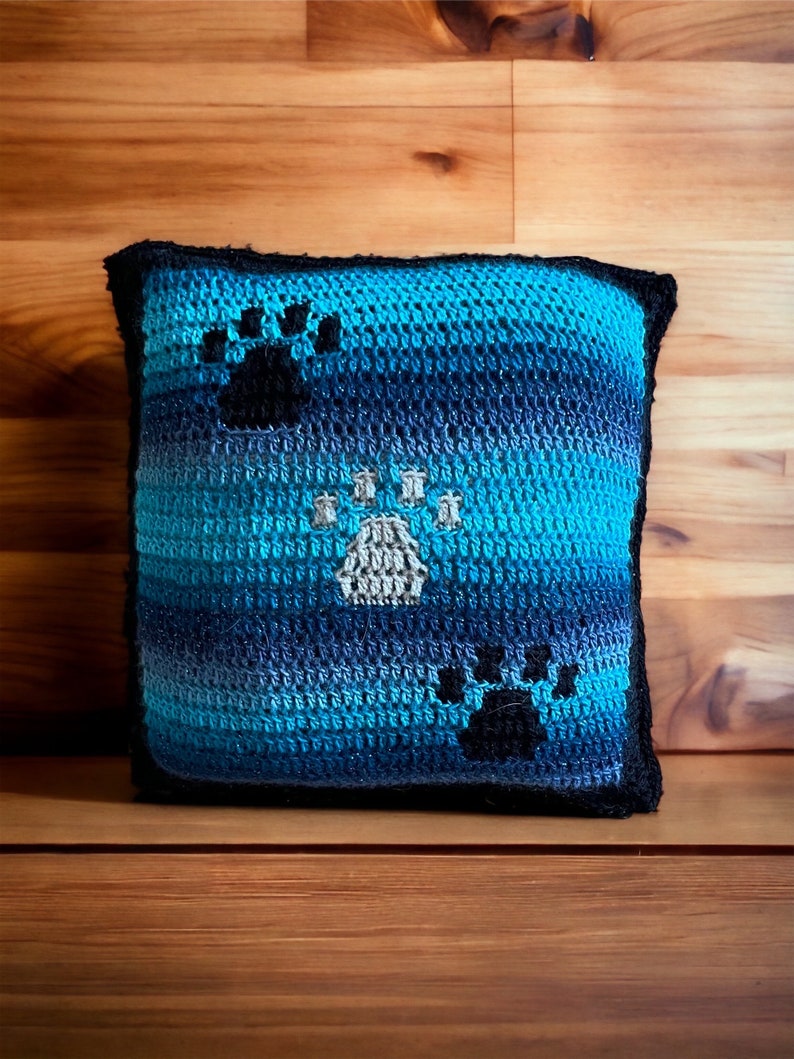 May include: A blue and black crocheted pillow with paw print designs. The pillow is made with a gradient yarn that transitions from dark blue to light blue.