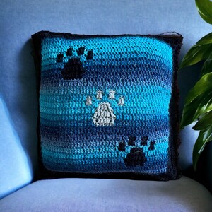 A blue and black crocheted cushion cover with three paw prints. The paw prints are in different shades of cream and black.