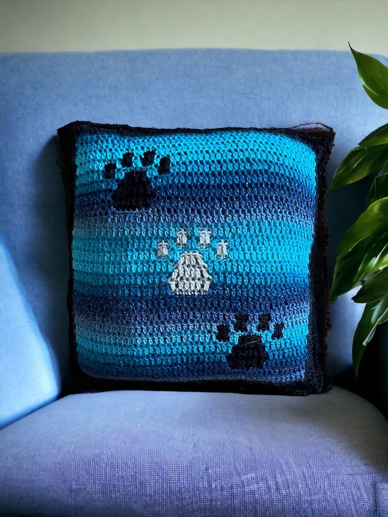 A blue and black crocheted cushion cover with three paw prints. The paw prints are in different shades of cream and black.