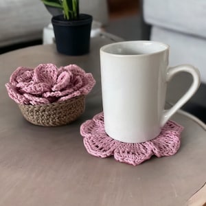 May include: A white ceramic mug with a handle sits on a pink crocheted coaster. A smaller pink crocheted coaster shaped like a flower is in the background.