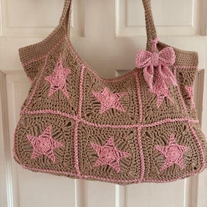 May include: A crocheted tote bag in a tan colour with pink star accents. The bag features a large, open design with a matching handle and a decorative pink bow. The bag is made of a textured yarn.
